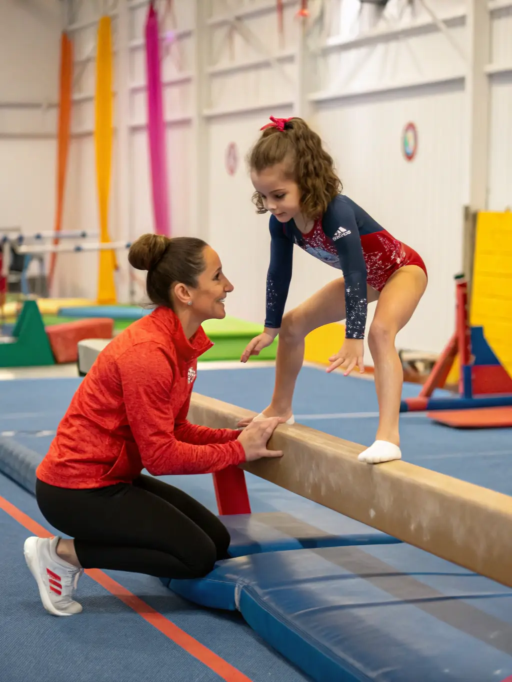 A photograph of a BBA GYM BOUC BEL AIR GYMNASTIQUE certified gymnastics coach spotting a young gymnast as she practices a back handspring on a padded mat. The coach is smiling and providing encouragement.